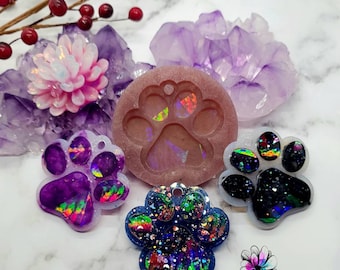 May include: A 2.25 inch silicone mold for making paw print charms. The mold is pink and has a paw print design. There are three finished charms made with the mold, one purple, one blue, and one black, all with iridescent glitter.