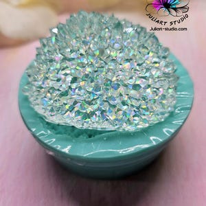May include: A teal-colored, round container holds a sparkling, crystal-like object. The crystals have a shimmering, iridescent quality. The container is set against a soft, blurred background. The logo for Juliart Studio is visible.