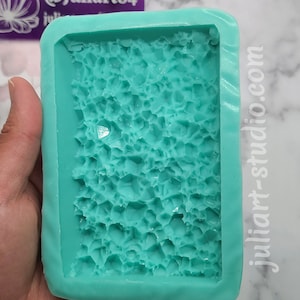 May include: A teal blue silicone mold with a textured surface resembling small rocks. The mold is rectangular and has a smooth, raised edge. The text "@Juliart84" and "juliart-studio.com" are visible on the mold.