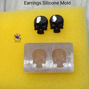 May include: A silicone mold for making skull-shaped earrings. The mold is silver and has two skull-shaped cavities. There are two black crystal skull earrings next to the mold.