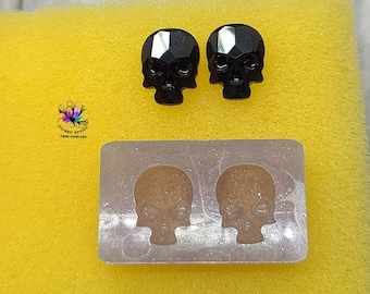 May include: A silicone mold for making skull-shaped earrings. The mold is silver and has two skull-shaped cavities. There are two black crystal skull earrings next to the mold.