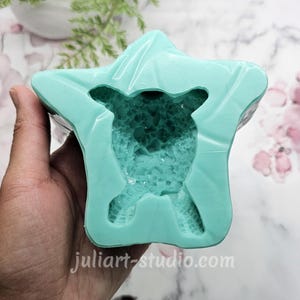 May include: A teal silicone mold in the shape of a star, with a detailed interior design. The mold is held in a hand, with the website address juliart-studio.com visible. The background features a marble pattern and greenery.