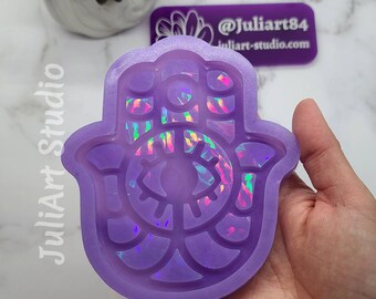 May include: A 4.75 inch purple silicone mold shaped like a Hamsa hand with a holographic eye design. The mold is for making resin coasters.