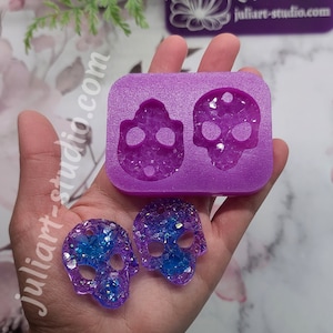 May include: Two purple resin skull earrings with blue glitter embedded in the resin. The earrings are shown next to a purple silicone mold of two skulls.