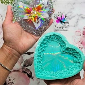 May include: A clear heart-shaped resin mold with a textured surface and a iridescent finish. The mold is being held in a hand, and a finished resin piece is also shown. The finished piece is a heart-shaped resin with a flower design and iridescent finish.