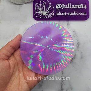 May include: A round, iridescent purple resin coaster with a swirling, rainbow-like pattern. The coaster is being held in a hand.