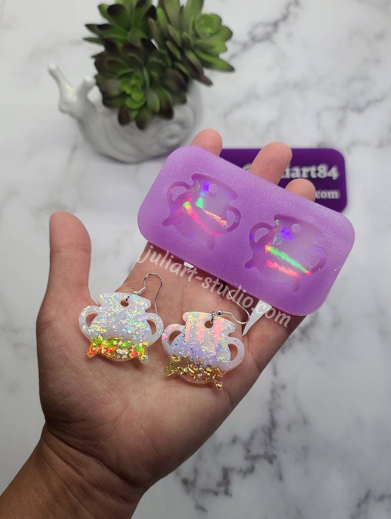 K&ouml;nnte beinhalten: Ein Paar regenbogenfarbene wei&szlig;e Harzohrringe in Form von Kesseln mit goldenen Glitzerakzenten. Die Ohrringe werden neben einer lila Silikonform mit zwei Kesselformen gezeigt.