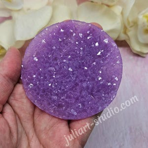 May include: A round, purple crystal geode with a sparkling surface is held in a hand. The geode is surrounded by white flowers and the text "juliart-studio.com".