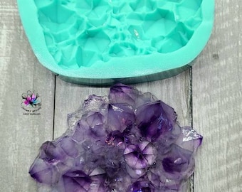 May include: A teal silicone mold shaped like a large crystal rock. The mold is on a light wood surface. A purple crystal rock is next to the mold.