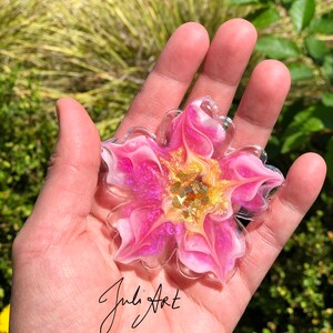 May include: A pink and white resin flower with gold glitter accents. The flower is shaped like a star and has a swirl design. The flower is about 4 inches in diameter.