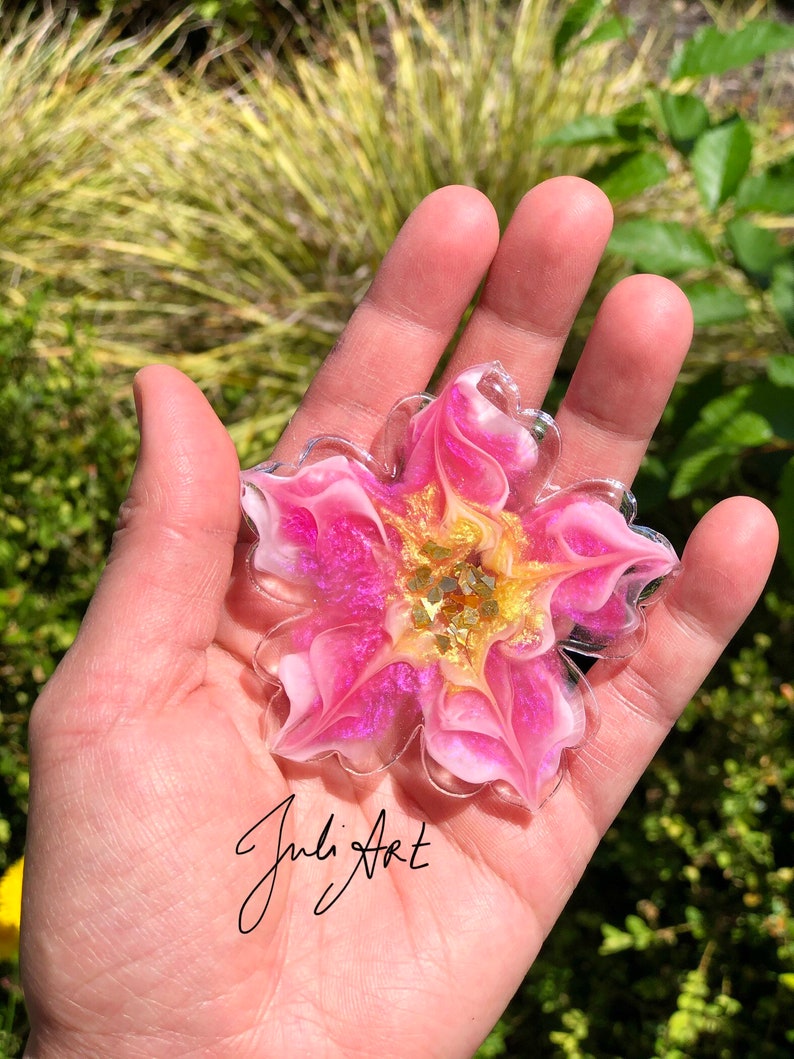 May include: A pink and white resin flower with gold glitter accents. The flower is shaped like a star and has a swirl design. The flower is about 4 inches in diameter.