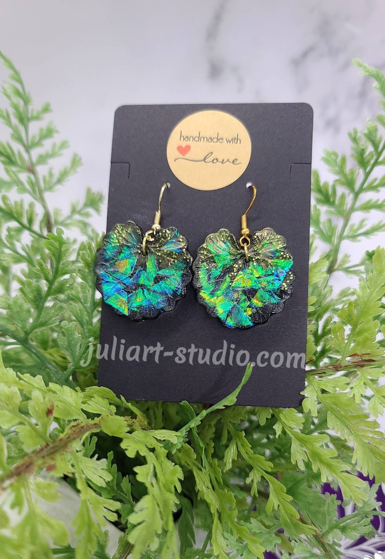 May include: A pair of iridescent green and black heart-shaped earrings with gold hooks. The earrings are handmade with love and are displayed on a black card with the text "handmade with love".