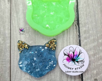 Könnte beinhalten: Eine grüne Silikonform in Form eines Katzenkopfes. Die Form ist mit klarem Glitter gefüllt. Neben der Form ist ein fertiges Harzstück zu sehen, das einen blauen und goldenen Katzenkopf mit Glitter zeigt.