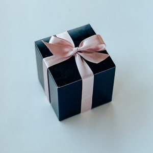 May include: A square black gift box featuring a smooth, matte finish. The box is wrapped with a light pink satin ribbon tied into a decorative bow on the top center. The ribbon extends vertically and horizontally across the sides of the box.