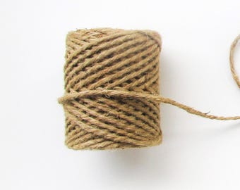 May include: A cylindrical spool of natural brown jute twine featuring a tightly wound, textured fiber construction. A single strand of the thin, fibrous cord extends outward from the center of the spool, showcasing the raw, earthy material.