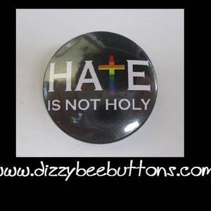 May include: A black button with a white cross and the text "HATE IS NOT HOLY". The cross is rainbow colored.