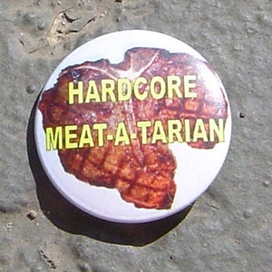 May include: A white button with a brown and red image of a meat cut. The button has the text "HARDCORE MEAT-A-TARIAN" in yellow letters.