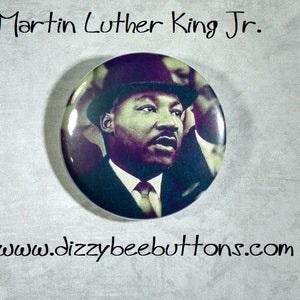 May include: A black and white button featuring a portrait of Martin Luther King Jr. wearing a hat and suit. The button has the text "Martin Luther King Jr." at the top and "www.dizzybeebuttons.com" at the bottom.