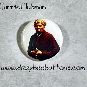 May include: A white button with a picture of Harriet Tubman. She is wearing a red dress and a yellow headscarf. The button has the text "Harriet Tubman" at the top and "www.dizzybeebuttons.com" at the bottom.