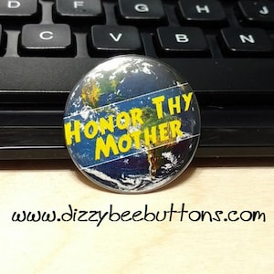 May include: A round button with a blue and green image of the Earth. The button has a yellow stripe across the middle with the text "HONOR THY MOTHER" in yellow letters.