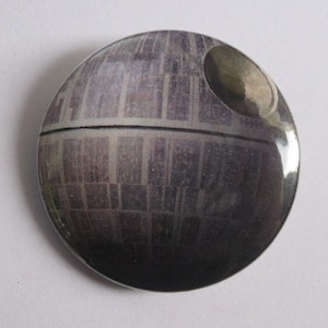 May include: A round, silver button with a black and white image of the Death Star from Star Wars.