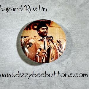 May include: A vintage button featuring a black and white photo of Bayard Rustin speaking at a podium with microphones. The button has a white background and the text "Bayard Rustin" is printed above the image.