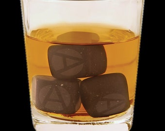 May include: A glass of amber-colored liquid with three dark gray whiskey stones. The stones have a raised letter "A" on each side.