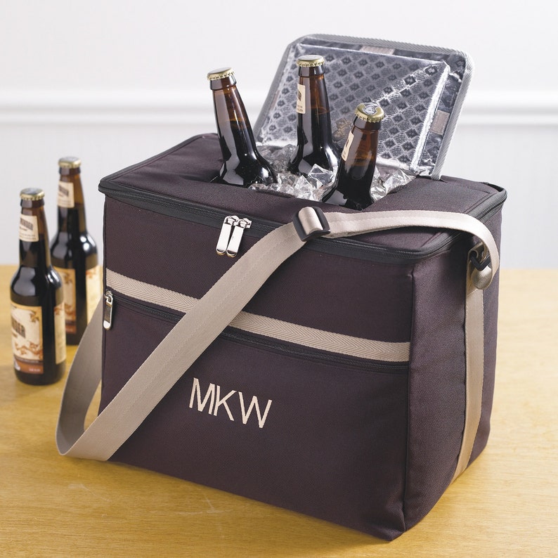 May include: A black insulated cooler bag with a tan shoulder strap and zipper closure. The bag has a white embroidered monogram "MKW" on the front. The cooler is filled with ice and bottles of beer.