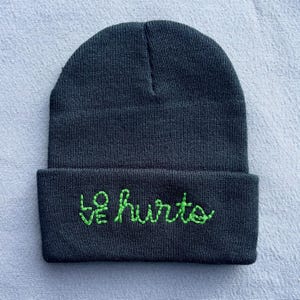 May include: Dark gray beanie with the embroidered words "love hurts" in bright green thread. The beanie is a classic, folded-brim style, perfect for cold weather. The text is in a cursive font.