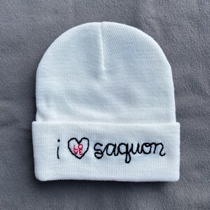 May include: White knit beanie with the embroidered text "i ❤️ saguon". The heart is red with the word "love" inside. The text is in a handwritten style. The beanie is folded at the brim.