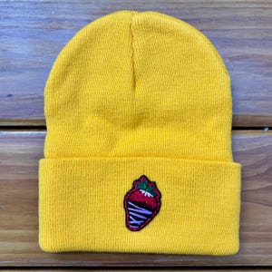 Puede incluir: Un gorro amarillo mostaza con un borde doblado. El gorro presenta un parche de una fresa cubierta de chocolate con una baya roja, hojas verdes y un glaseado morado. El gorro está hecho de material de punto.