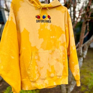 May include: A yellow hoodie with a bleached design and a front pocket. The hoodie features the word "DAYDREAMER" embroidered below a design of three embroidered fruits. The hoodie is hanging outside on a tree branch.