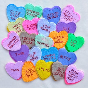 Puede incluir: Una colección de decoraciones en forma de corazón de fieltro colorido con texto bordado. Los corazones presentan frases como "LOVE ALWAYS" y "BESTIE". Cada corazón tiene un color diferente y un borde contrastante.