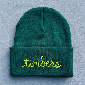 May include: A dark green knit beanie with the word "timbers" embroidered in a bright yellow cursive font. The beanie is folded at the brim and sits on a light blue surface. This winter hat is a stylish accessory.