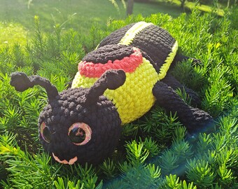 May include: A crocheted plush toy shaped like a bumblebee with black and yellow stripes, red accents, and large googly eyes. The toy is lying on a bed of green foliage.