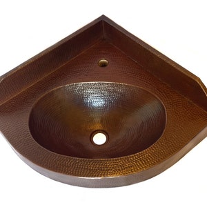 Puede incluir: Un lavabo de esquina de cobre con acabado martillado. El lavabo es de forma ovalada y tiene un agujero de drenaje en el centro.