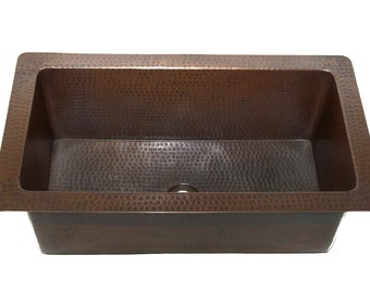 Puede incluir: Un fregadero de cocina rectangular de cobre con acabado martillado. El fregadero tiene un solo agujero de drenaje en el centro.
