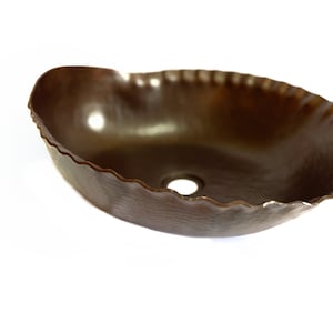 May include: A brown, oval-shaped, hammered copper vessel sink with a scalloped edge. The sink has a single drain hole in the center.