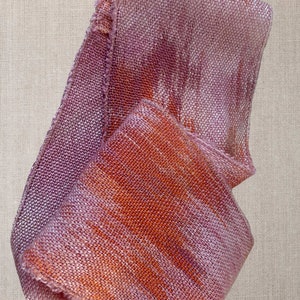 May include: A soft, woven scarf in shades of pink and orange with a subtle shimmer. The scarf is folded and draped on a textured surface.