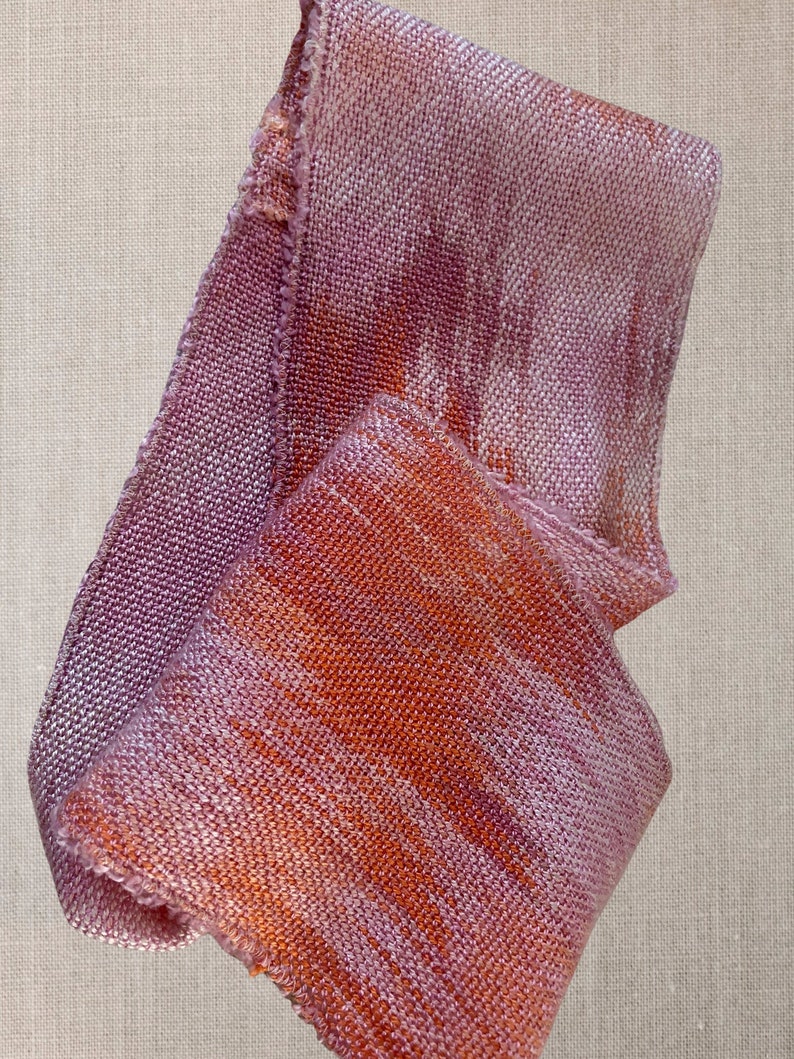 May include: A soft, woven scarf in shades of pink and orange with a subtle shimmer. The scarf is folded and draped on a textured surface.