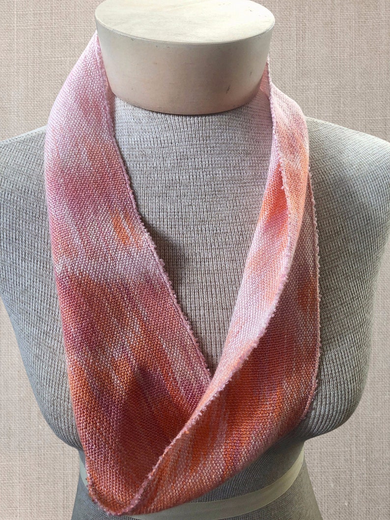 May include: A pink and orange woven fabric neck scarf. The scarf is folded in half and draped over a mannequin's neck.
