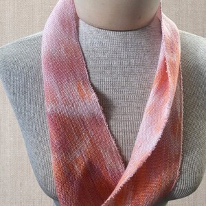 May include: A pink and orange woven fabric scarf with a textured pattern. The scarf is displayed on a mannequin.