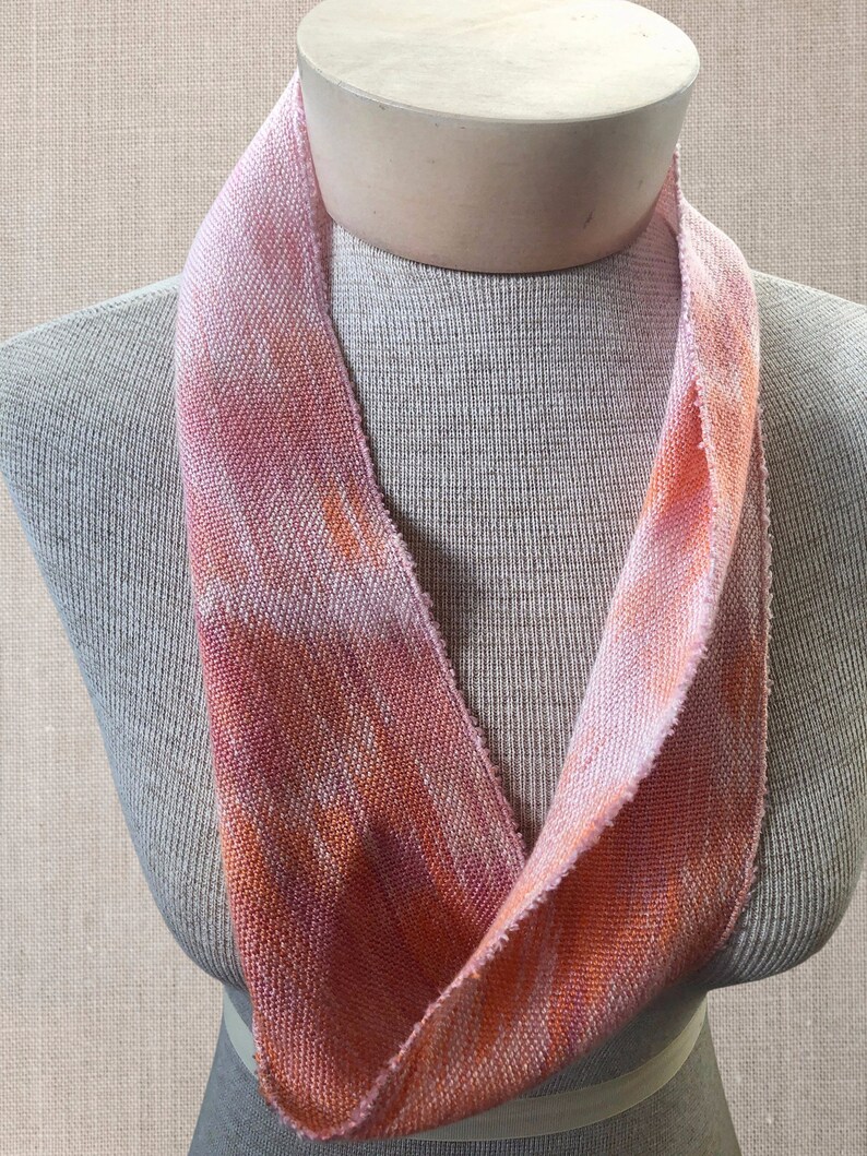 May include: A pink and orange woven fabric scarf with a textured pattern. The scarf is displayed on a mannequin.