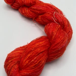 May include: A vibrant skein of hand-dyed yarn in shades of red and orange. The yarn appears to be a blend of fibers, with a textured, slightly irregular appearance. The colors blend and shift, creating visual interest. Ideal for knitting or crochet projects.
