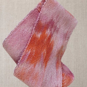 May include: A pink and orange woven fabric with a textured surface. The fabric is folded and has a frayed edge.