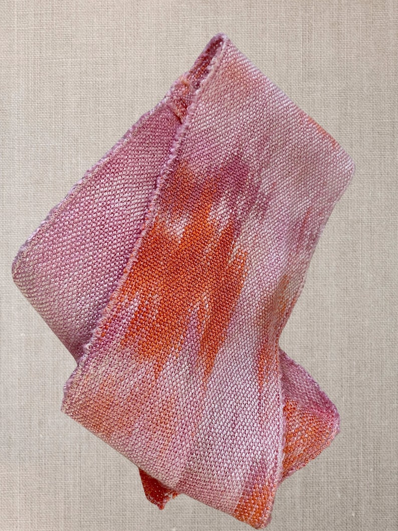 May include: A pink and orange woven fabric with a textured surface. The fabric is folded and has a frayed edge.