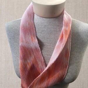 May include: A pink and orange woven scarf with a tie-dye pattern. The scarf is displayed on a mannequin.