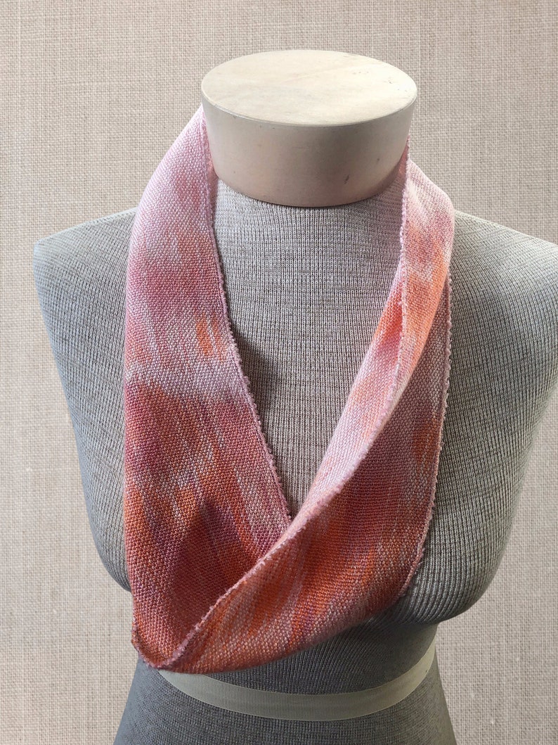 May include: A pink and orange woven scarf with a tie-dye pattern. The scarf is displayed on a mannequin.