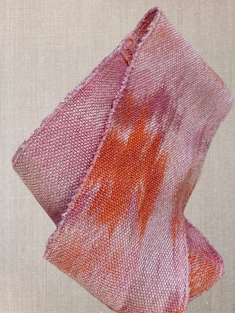 May include: A pink and orange woven fabric with a textured surface. The fabric is folded in half and has a frayed edge.