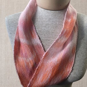 May include: A pink and orange woven scarf with a tie-dye pattern. The scarf is displayed on a mannequin.